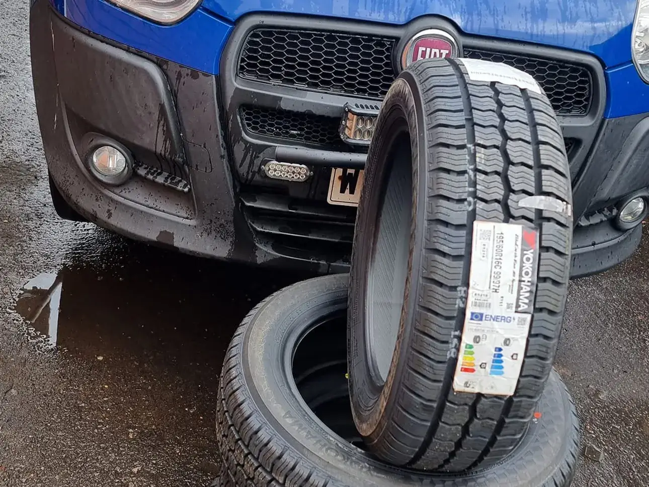 Two new replacement tyres ready to be fitted beside a car.